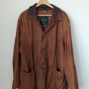 May include: A brown leather jacket with a collar and two front pockets hangs from a wooden hanger. The jacket has a button-up front and a label that reads "DANIER CANADA".