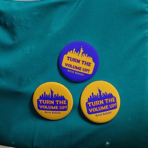 May include: A teal tote bag with black handles and a gray zipper pocket. Three round buttons are pinned to the bag, each with the text "TURN THE VOLUME UP!" and a skyline graphic in yellow and blue.
