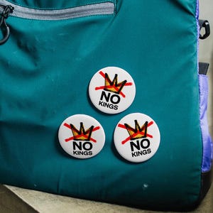 Puede incluir: Bolso tote de color turquesa con asas negras y un bolsillo con cremallera gris. Tres botones blancos con un diseño de corona roja y dorada y el texto "NO KINGS" están prendidos en la bolsa. La bolsa tiene detalles morados.