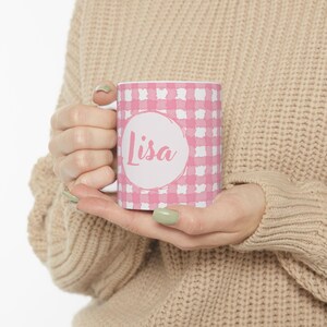 May include: A white ceramic mug with a pink gingham pattern. The mug features a white circle with the name "Lisa" in pink script. The mug is held in a person's hands.