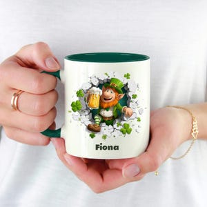 May include: White ceramic mug with a green interior and handle. The mug features a cartoon leprechaun holding a beer, emerging from a cracked wall, surrounded by shamrocks. The name "Fiona" is printed on the mug.