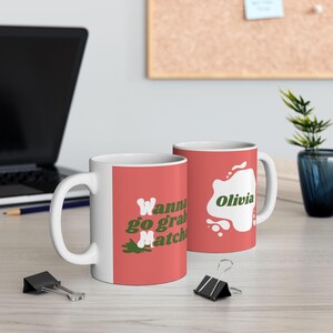 May include: Two white ceramic mugs with a red background. One mug has the text "Wanna go grab Matcha" in white and green. The other mug has the name "Olivia" in green. Both mugs are on a light-coloured surface.