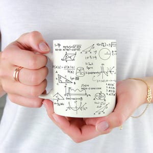 Może przedstawiać: Biały ceramiczny kubek pokryty czarnymi wzorami i równaniami matematycznymi. Kubek zawiera różne kształty geometryczne, funkcje trygonometryczne i wyrażenia algebraiczne, tworząc motyw naukowy i edukacyjny.