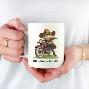 Könnte beinhalten: Weißer Keramikbecher mit einem Cartoon-Frosch in einem Cowboyhut, der ein rotes Motorrad fährt. Der Frosch trägt ein rotes Halstuch und eine braune Ledertasche. Der Text "Off to Cause a Kerfuffle" ist darunter gedruckt.