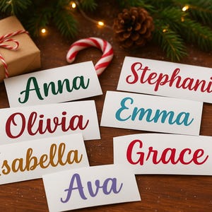 Op de afbeelding: Een kersttafereel met naamkaartjes met namen als Anna, Olivia en Stephanie in verschillende kleuren. Een ingepakt cadeau, een zuurstok, dennentakken en lichtjes maken de feestelijke scène compleet.