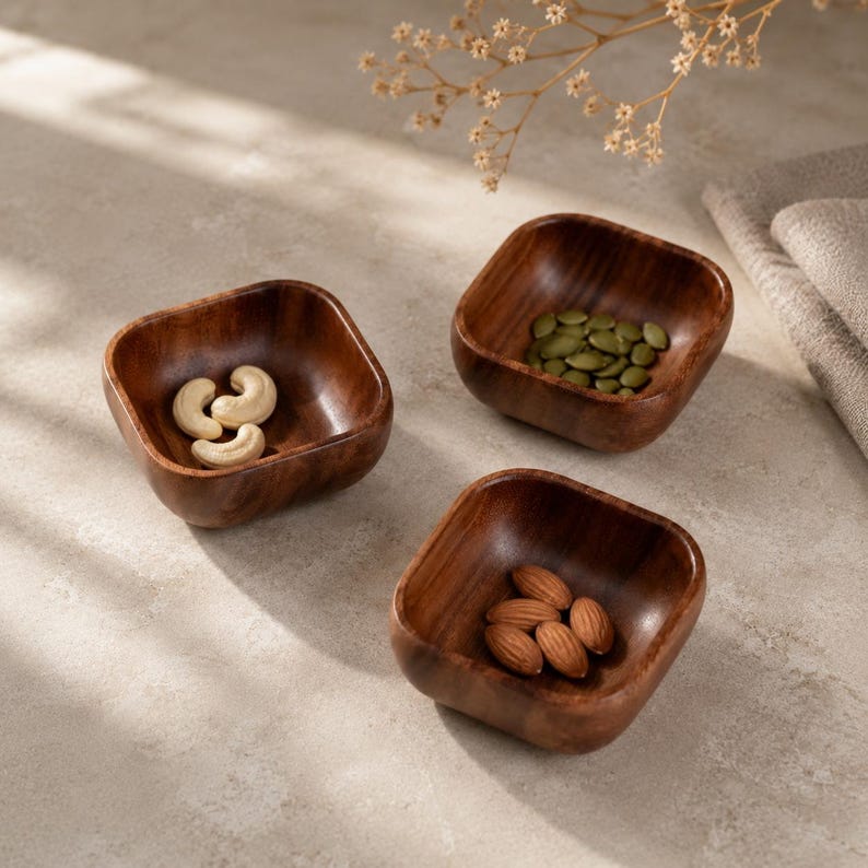 May include: Three small, square-shaped wooden bowls with rounded corners and a dark brown wood grain finish. One bowl contains raw cashews, another holds green pumpkin seeds, and the third bowl is filled with whole brown almonds.