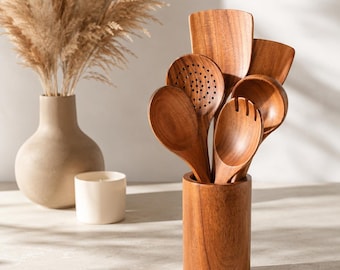 May include: A set of brown wooden kitchen utensils including a slotted spoon, a solid spoon, a spatula, and a fork, all stored upright in a cylindrical wooden holder. The utensils feature a smooth finish and visible wood grain throughout the set.