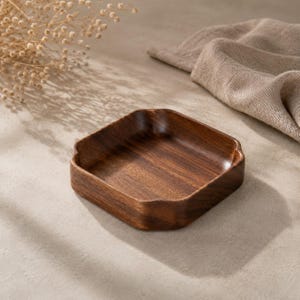 May include: A square wooden tray with rounded corners and a deep, dark brown wood grain finish. The tray features a smooth, polished surface with slightly raised edges, showcasing the natural texture and rich color variations of the wood.