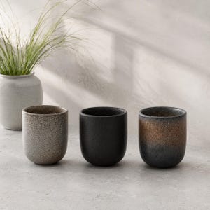 May include: Three cylindrical ceramic cups with rounded bases. The left cup has a speckled light beige finish, the center cup is solid matte black, and the right cup features a gradient transition from dark charcoal at the base to a warm bronze top.