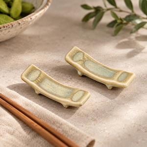 May include: Two curved ceramic chopstick rests with a glossy, mottled light green and beige glaze. Each rest features a slightly concave center and small feet on the underside to elevate the chopsticks from the table surface.