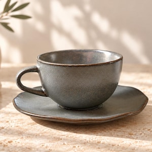 May include: A ceramic teacup and matching saucer set in a dark, mottled gray glaze with a smooth, rounded handle. The cup features a deep basin, while the saucer has a slightly irregular, organic edge, both showcasing a subtle, reflective finish.