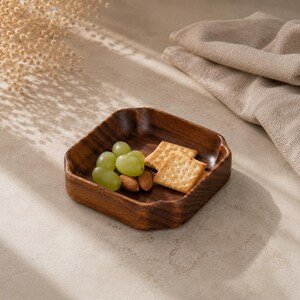 May include: A square wooden bowl with rounded corners and a dark brown wood grain finish. The bowl contains a small cluster of green grapes, two light brown almonds, and two square, light-colored crackers with small holes on their surface.
