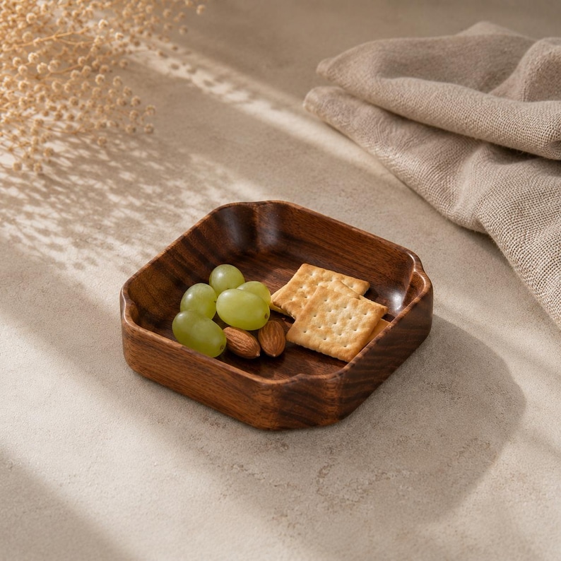 May include: A square wooden bowl with rounded corners and a dark brown wood grain finish. The bowl contains a small cluster of green grapes, two light brown almonds, and two square, light-colored crackers with small holes on their surface.