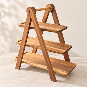 May include: A three-tiered wooden serving stand featuring a collapsible A-frame design. The structure consists of two triangular side supports connected by a top handle, holding three rectangular wooden trays with smooth, natural grain finishes.