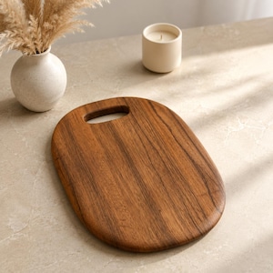 May include: An oval-shaped wooden cutting board with a smooth, natural brown grain and a cutout handle at the top. The board features a warm, polished finish, showcasing the distinct vertical wood grain patterns across its flat, solid surface.