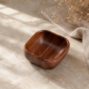 May include: A small, square-shaped wooden bowl with rounded corners and a smooth, polished finish. The bowl features a rich, dark brown wood grain pattern throughout its interior and exterior surfaces, showcasing a deep, natural color variation.