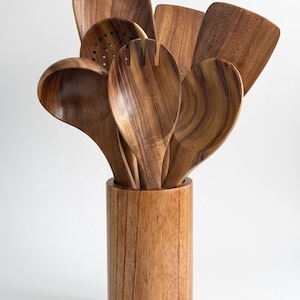 May include: A collection of wooden kitchen utensils, including a slotted spoon, spatula, and serving spoons, displayed in a cylindrical wooden holder. The utensils and holder are made of a light brown wood with visible grain.