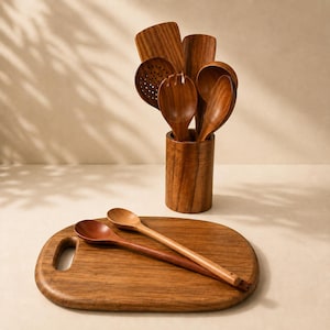 May include: A set of dark brown wooden kitchen utensils including a slotted spoon, a spatula, a fork, and round spoons stored in a cylindrical wooden holder, with two additional wooden spoons resting on an oval wooden cutting board.