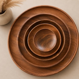 May include: A set of five circular wooden plates or shallow bowls of varying sizes, nested one inside the other. Each piece features a smooth, polished finish with visible natural wood grain patterns in shades of medium to dark brown.
