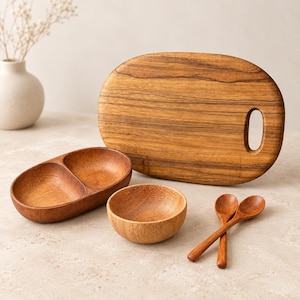 May include: A set of wooden kitchenware including an oval cutting board with a handle hole, a dual-compartment oval serving dish, a small round bowl, and two matching wooden spoons, all featuring a natural brown wood grain finish.