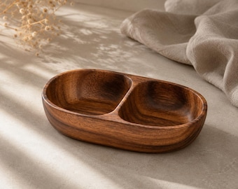 May include: An oval-shaped wooden serving dish featuring two distinct, side-by-side compartments. The dish is crafted from dark brown wood with a visible, natural grain pattern throughout the smooth, curved interior and exterior surfaces.