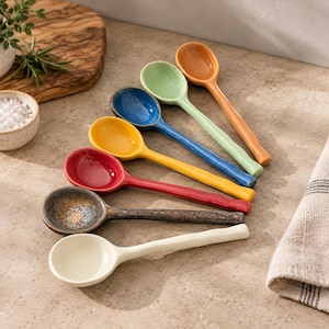 May include: A set of seven ceramic spoons with long, slightly wavy handles, arranged in a diagonal row. The spoons feature a variety of glossy finishes in solid colors including cream, dark brown, red, yellow, blue, light green, and orange.