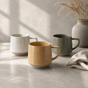 May include: Three ceramic mugs with handles, each featuring a speckled finish and a smooth base. One mug is white with dark speckles, one is a warm mustard yellow with brown speckles, and one is a muted sage green with a solid, smooth texture.