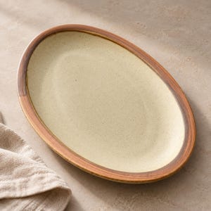 May include: An oval ceramic serving platter featuring a speckled cream-colored center and a wide, dark brown rim with a visible brushstroke texture. The plate has a smooth, slightly concave surface and a distinct border that frames the interior.