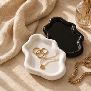 May include: Two wavy-edged ceramic trinket dishes, one in glossy white and one in glossy black. The white dish holds three gold-toned rings and a delicate gold chain necklace with a small circular pendant. The items are arranged for display.