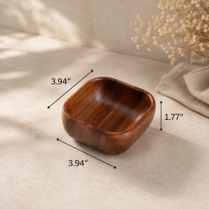 May include: A square wooden bowl with rounded corners and a smooth, dark brown grain finish. The bowl features dimensions of 3.94 inches in length, 3.94 inches in width, and 1.77 inches in height as indicated by the visible measurement labels.