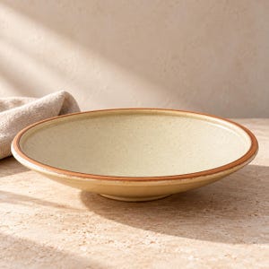 May include: A shallow, wide ceramic bowl with a speckled cream-colored interior and a smooth, dark brown rim. The bowl features a low-profile design with a slightly flared edge, showcasing a natural, earthy aesthetic with a subtle, textured finish.