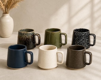 Puede incluir: Una colección de seis tazas de cerámica con diversos acabados, incluyendo azul oscuro, crema moteado, marrón mate, negro con motas, verde oliva sólido y una taza con degradado marrón y azul, todas con un asa resistente.