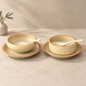 May include: Two sets of ceramic tableware, each consisting of a round bowl and a matching plate with a speckled cream finish and a burnt orange rim. Each bowl contains a cream-colored ceramic spoon resting inside, completing the dining set.