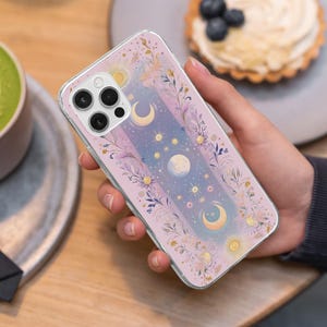 May include: A clear phone case with a celestial design. The case features moons, stars, and suns in pastel colours, with a floral border and a soft pink background. The phone case is held in a person's hand.