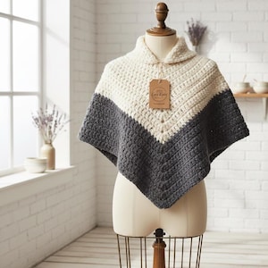 Könnte beinhalten: Ein handgefertigter Häkelponcho in Creme und Anthrazitgrau, ausgestellt auf einer Schaufensterpuppe. Der Poncho hat ein V-förmiges Farbblockdesign und einen Kragen. Ein Etikett mit dem Text "Cozy Knits" hängt am Kragen.