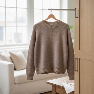 May include: A neutral-toned, crew-neck sweater hangs on a wooden hanger. The sweater has a textured knit pattern with flecks of color throughout. The sleeves are long, and the hem and cuffs are ribbed. The sweater is displayed indoors.