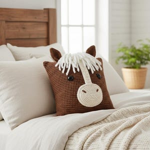 May include: A brown crochet horse-shaped pillow with a white mane and muzzle, black button eyes, and brown ears. The pillow is placed on a bed with white bedding and a cream-colored blanket. The pillow is a decorative item.