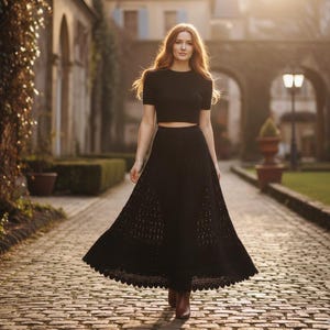 Puede incluir: Un conjunto negro de dos piezas que consta de un top corto de manga corta y una falda larga y fluida con dobladillo festoneado. El conjunto se lleva con botas marrones. La mujer camina por un camino adoquinado.