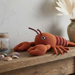 May include: A crocheted orange lobster with dark red stripes, lying on a wooden surface. The lobster has large claws and a detailed design. Decorative elements include seashells, a vase with dried plants, and glass jars.