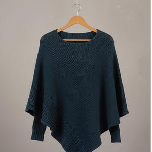 Puede incluir: Un poncho tejido de color verde azulado con cuello redondo y mangas largas. El poncho está colgado de una percha de madera sobre un fondo neutro. Las mangas tienen un patrón texturizado.