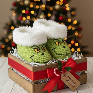 May include: A pair of green crocheted baby booties with a white fluffy trim, featuring a cartoon face design. The booties are presented in a wooden crate with a red ribbon and a tag that says "Handmade with Love!"