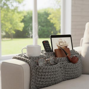 Può includere: Un organizer per bracciolo del divano grigio lavorato all'uncinetto con più tasche. Contiene una tazza bianca fumante, un telecomando, un tablet che mostra una nave, un portafoglio in pelle marrone, una pipa e occhiali. L'organizer è su un bracciolo del divano beige.