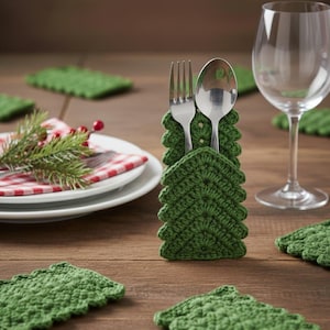 May include: A green crocheted Christmas tree-shaped utensil holder with a fork and spoon inside. A white plate with a red checkered napkin and a sprig of pine is in the background. Several green tree-shaped coasters are also visible.
