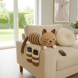 May include: A crocheted cat-shaped armrest organizer in tan and brown, with a striped body and a smiling face. It features two pockets for holding items and is resting on a beige sofa arm. A remote control is also visible.
