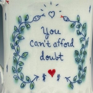 May include: White ceramic mug with a blue and green floral design. The mug features the text "You can't afford doubt" in blue script, along with a red heart and dollar signs.