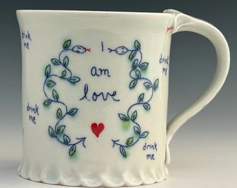 Puede incluir: Taza de cerámica blanca con borde ondulado y asa curva. La taza está decorada con un diseño de vid azul y verde, dos ilustraciones de serpientes, las palabras "I am love", un corazón rojo y las palabras "drink me".