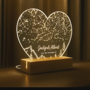 May include: Heart-shaped night light with a constellation design. The clear acrylic heart is engraved with stars, mountains, and trees, and rests on a wooden base. The names "Jaclyn & Albert" and date are also engraved.