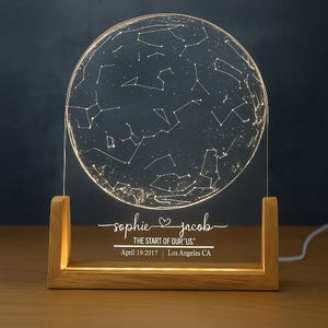May include: A personalized night light featuring a clear, circular panel etched with a constellation map. The light sits on a wooden base, with the names "sophie & jacob" and the text "THE START OF OUR US" and date and location printed below.