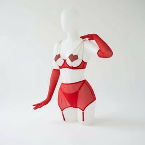 May include: Red lingerie set on a mannequin. The set includes a bra with heart-shaped embellishments, a high-waisted garter belt, and matching red gloves. The bra features gold chain straps. The lingerie is displayed against a white background.
