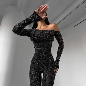 May include: Black jumpsuit with long sleeves and an off-the-shoulder neckline. The jumpsuit is embellished with small, sparkling details and features cut-out patterns on the sleeves and waist. The lower legs have a sheer lace design.